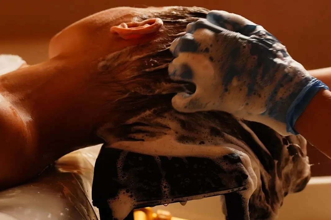 Close-up of a head spa therapist's gloved hands massaging cleansing foam into a reclining client's scalp at the wash basin, viewed from behind the crown